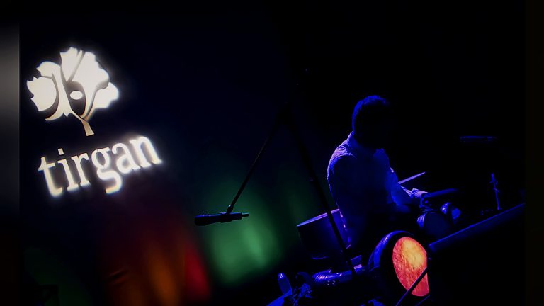 Musician playing in blue light beside the Tirgan logo, Tirgan Nowruz Festival 2018